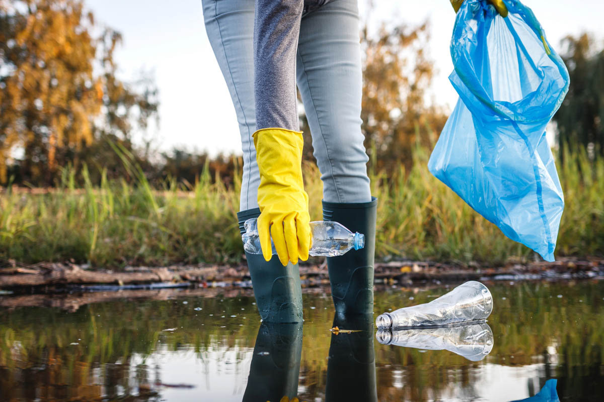 A retention pond clean up expert in rubber gloves is holding a plastic bag in a pond.