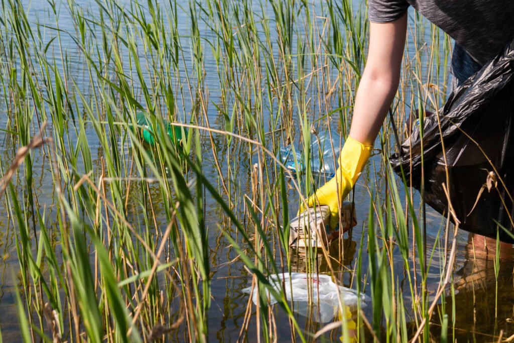 A woman providing retention pond maintenance wearing a yellow glove is picking up trash in the water.