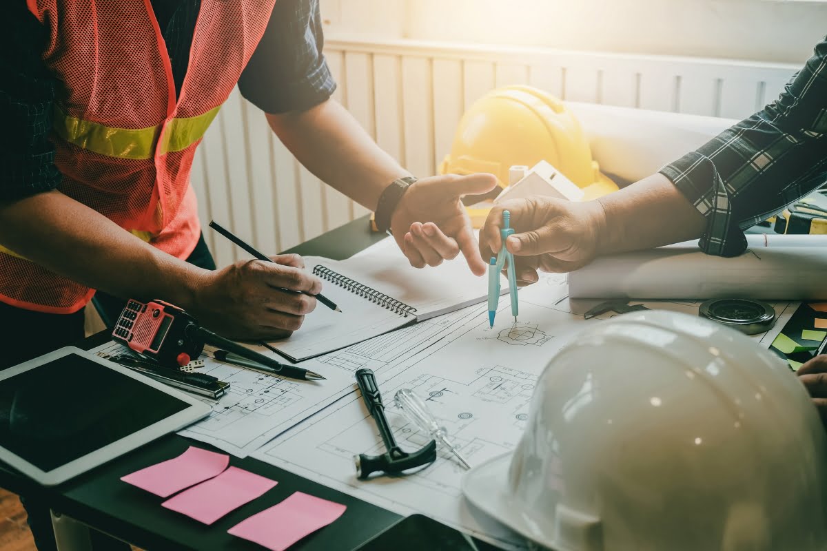 Two individuals discussing a building project over a table filled with blueprints, notepad, helmet, and construction tools. One person is using a compass while the other writes in the notepad, planning every detail of the land clearing process.