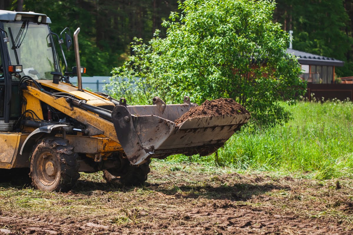 A yellow construction vehicle with a dirt-filled bucket, used for land clearing, is parked on a grassy area with a tree and building in the background.