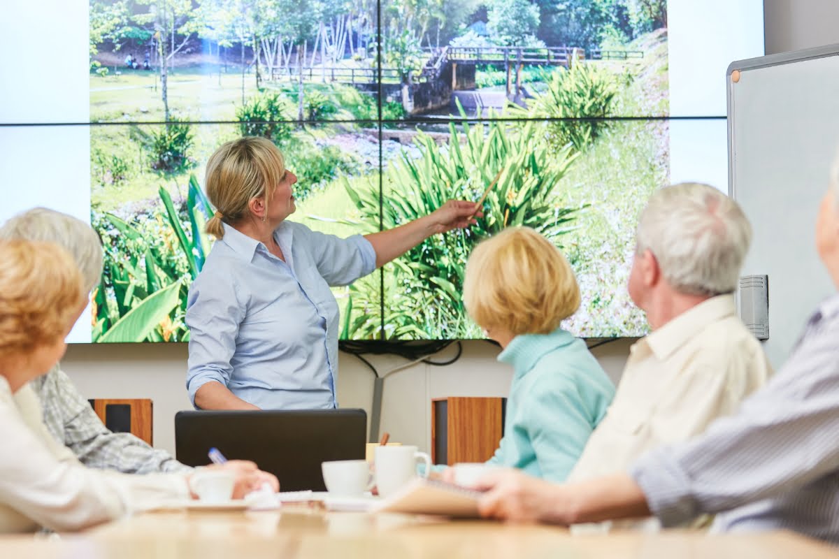 A person is giving a presentation to a group of seated individuals, pointing at a large screen displaying a lush, outdoor landscape. They discuss the impact of land clearing on natural habitats and the importance of sustainable practices.