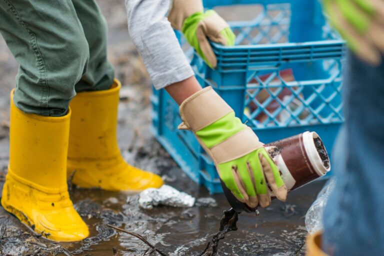 Two people wearing gloves and yellow boots, part of a storm cleanup company, are cleaning up muddy ground; one is placing a muddy jar into a blue crate.