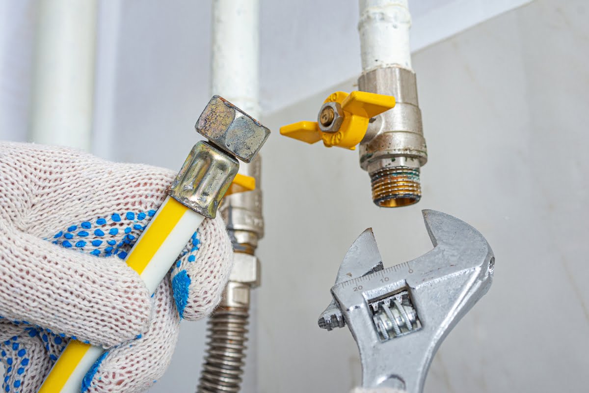 A gloved hand holds a yellow gas hose, preparing to connect it to a pipe with an adjustable wrench near a shut-off valve with a yellow handle, showing what to do after a storm for safe utility reconnection.