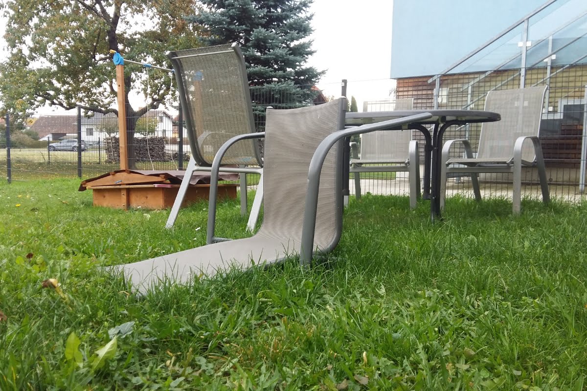 Outdoor scene with a table and several chairs on a grassy lawn. One chair has a broken seat, causing the fabric to hang down to the ground, much like what to do after a storm when assessing damage. Trees and a building are visible in the background.