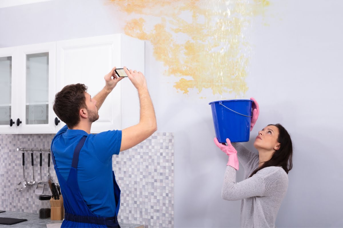 A woman holds a blue bucket to catch leaking water from the ceiling, showing what to do after a storm, while a man in a blue shirt takes a photo of the leak in the kitchen.