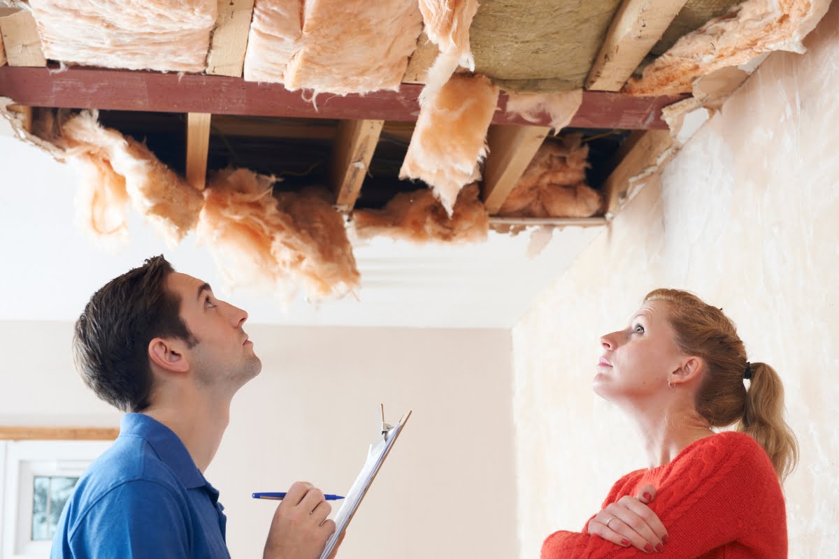 A man with a clipboard and a woman look up at exposed insulation and a damaged ceiling in a house, pondering what to do after a storm.