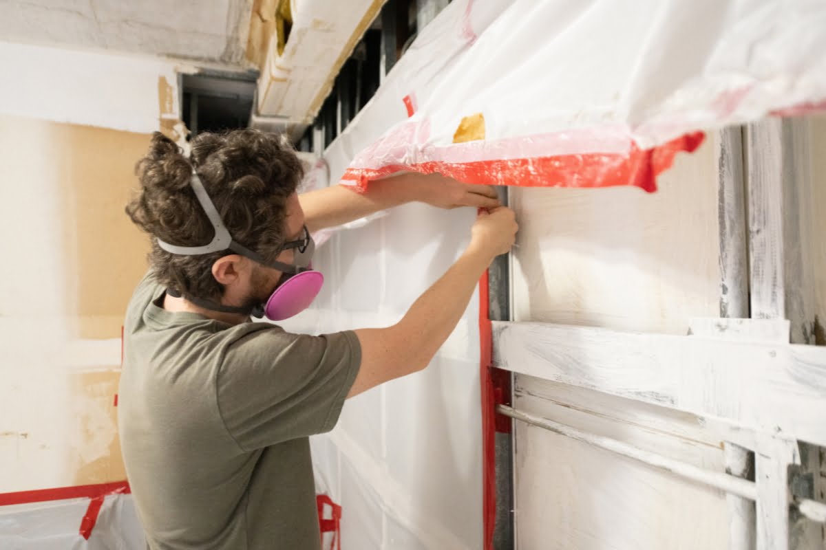 After a storm, a person wearing safety goggles, a respirator, and a green shirt is carefully applying red tape to white plastic sheeting on a wall in a construction or renovation setting.