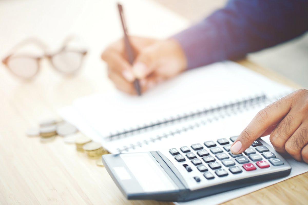 A person is using a calculator and writing in a notebook at a wooden table. Nearby are coins, a pair of eyeglasses, and brochures about professional tree removal.