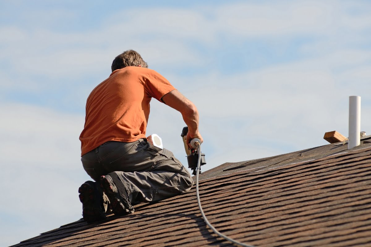 A person in an orange shirt is kneeling on a roof under a blue sky, using a nail gun to attach shingles and storm proofing your home.