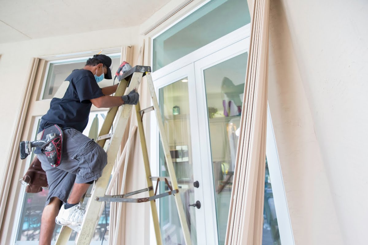 A man on a ladder installs wooden trim around large windows and a sliding glass door in a residential space, focusing on storm proofing your home.