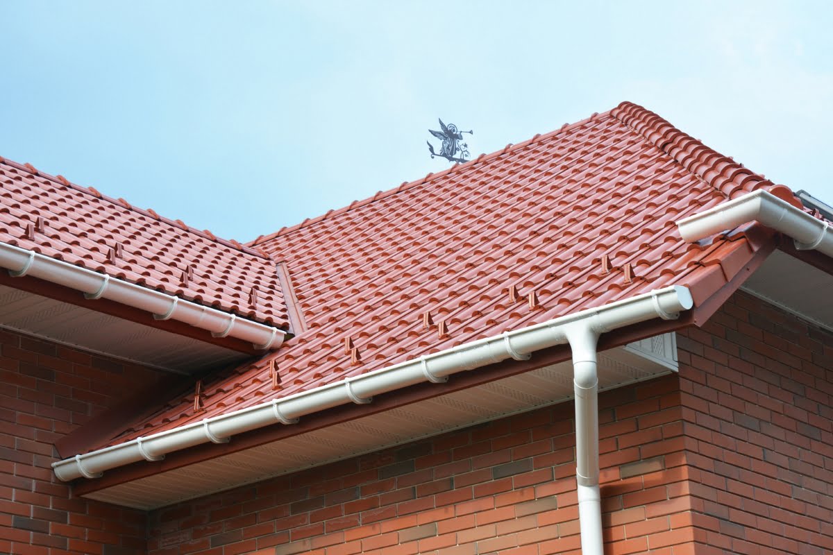 A red tiled roof with white gutters on a brick building against a blue sky, featuring a weather vane at the peak—an ideal design for storm proofing your home.