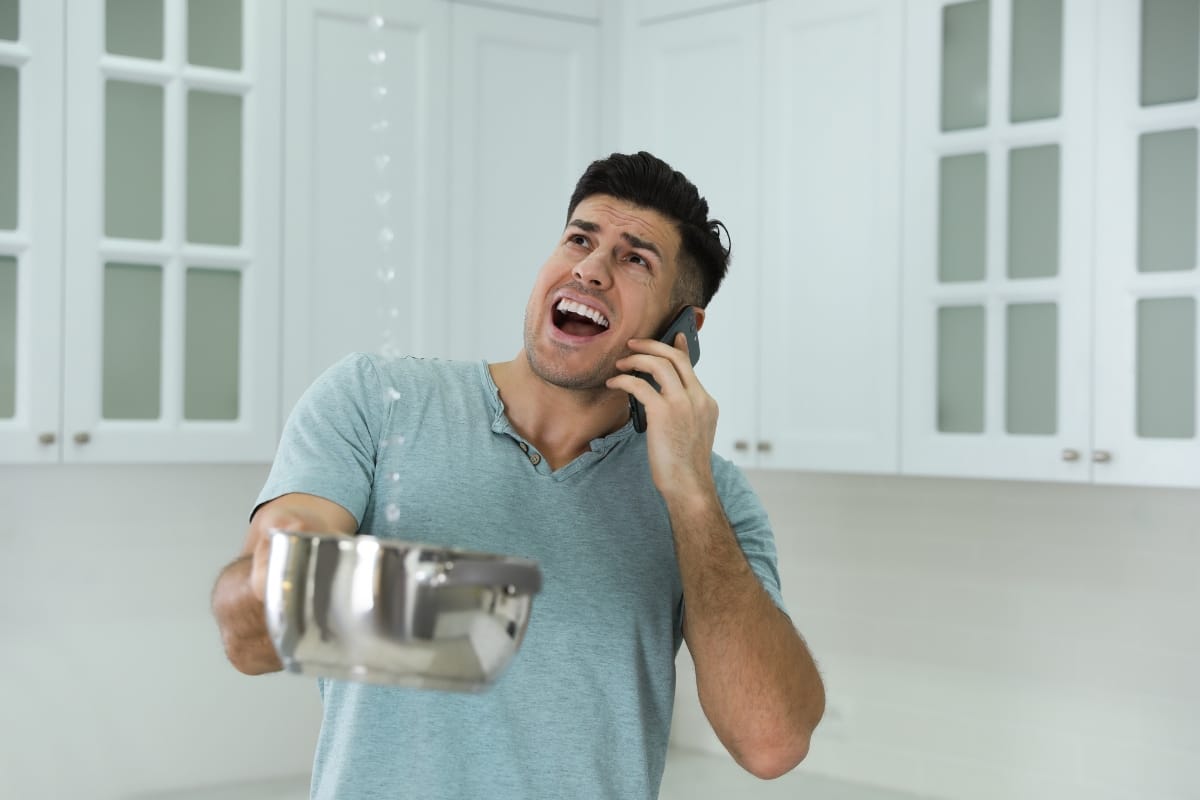 Person holding a pot to catch leaking water from the ceiling, while negotiating storm damage insurance claims on the phone in a kitchen with white cabinets.