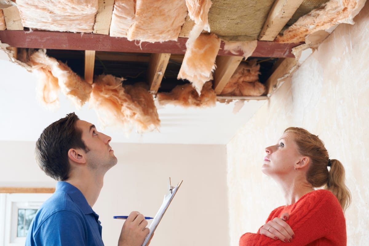 Two people look up at a ceiling with exposed insulation and wooden beams, assessing the scene for storm damage. One holds a clipboard for taking notes on the insurance claims, while the other stands with arms crossed, deep in thought.