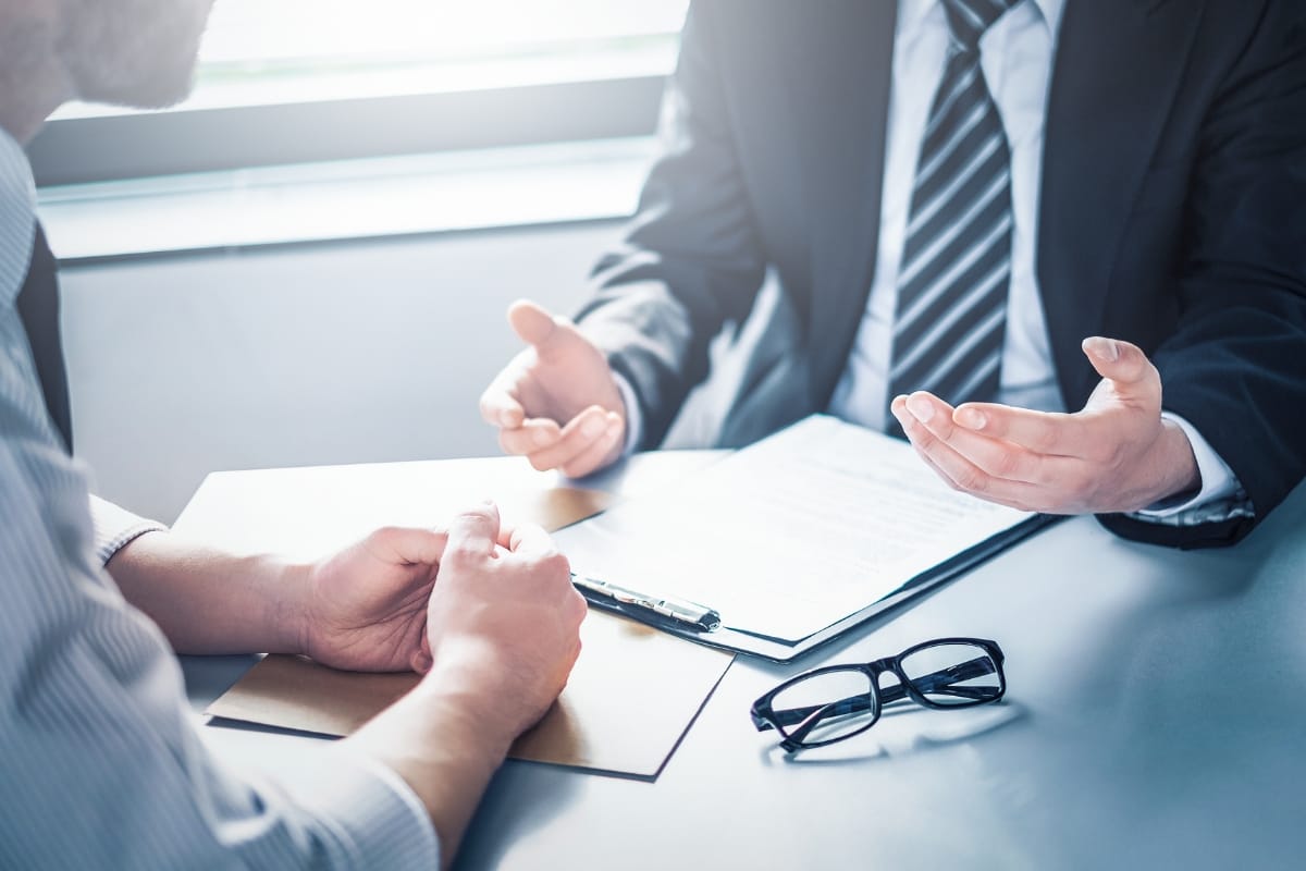 Two people in formal attire are seated at a table, discussing storm damage insurance claims. A clipboard with papers and a pair of glasses are on the table.