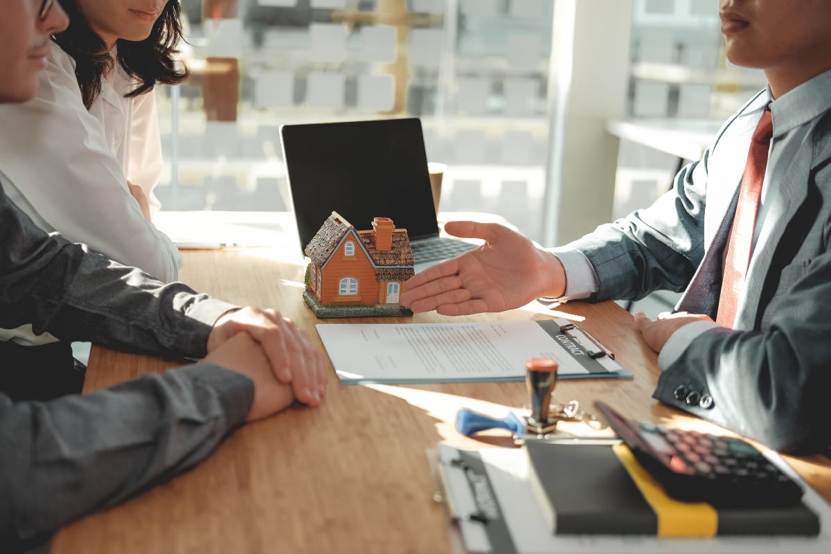 In an office setting, people sit at a table engaged in discussing storm damage insurance claims, surrounded by documents and a small house model.