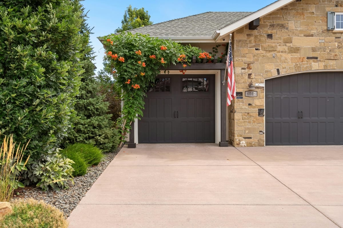 A stone house features a two-car garage with dark doors, proudly displaying an American flag. Above the left door, green foliage with orange flowers thrives, hinting at potential tree root damage nearby.