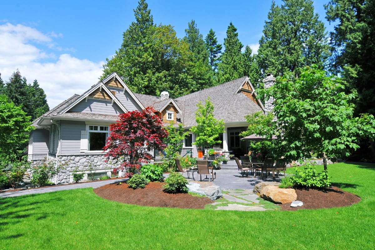Single-story gray house with stone accents, surrounded by lush green lawn, best trees, and landscaped garden beds under a partly cloudy sky. Outdoor seating area visible near the entrance.