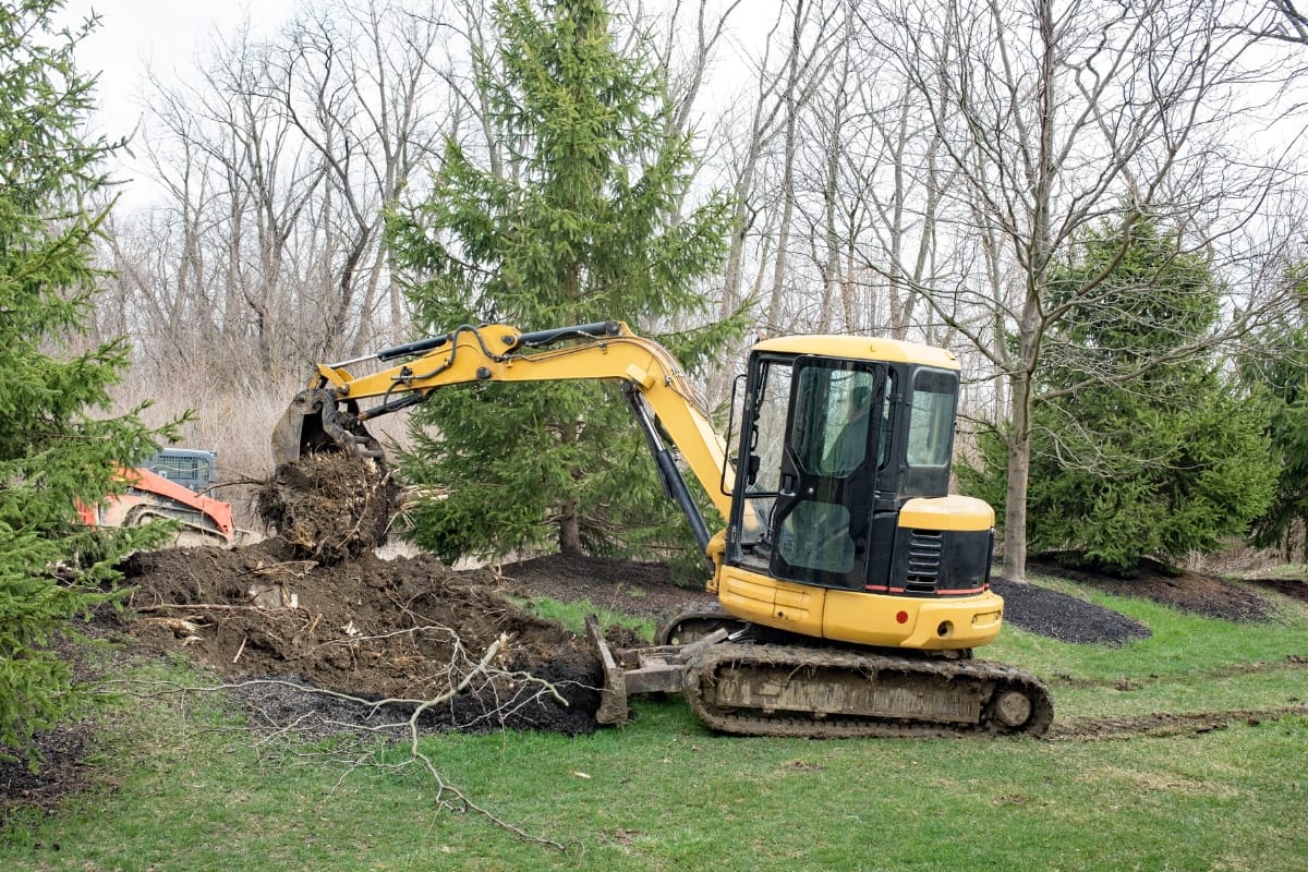 A yellow excavator is digging up soil and debris in a grassy area surrounded by trees on a cloudy day, assisting with hazardous tree removal.