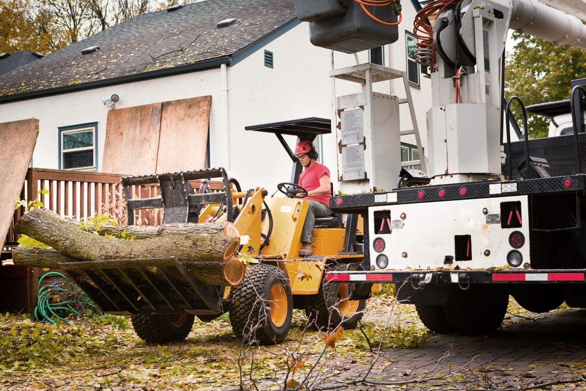 A worker performs hazardous tree removal, operating heavy machinery to move a large fallen trunk next to a house with boarded windows and scattered debris.